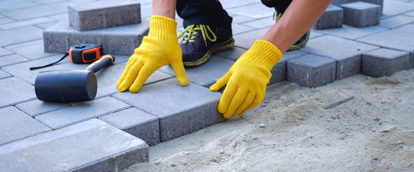 The master in yellow gloves lays paving stones in layers. Garden brick pathway paving by professional paver worker. Laying gray concrete paving slabs in house courtyard on sand foundation base. S